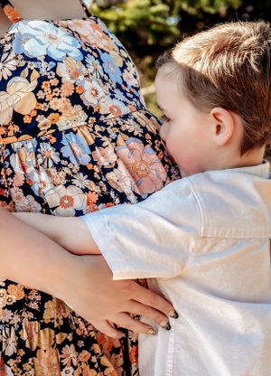 A tender moment as a child embraces a floral dress-clad figure in a sun-dappled outdoor setting.