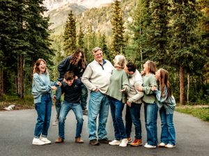 A group poses in a playful line formation on a mountain road surrounded by tall pine trees.