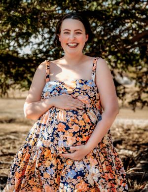 A floral maternity dress flows elegantly in warm sunlight against a natural backdrop.