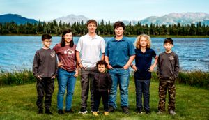 A group stands together by a lake with mountains and evergreen forest visible in the background.
