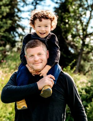 A joyful outdoor portrait showing a child riding on someone's shoulders against a nature backdrop.