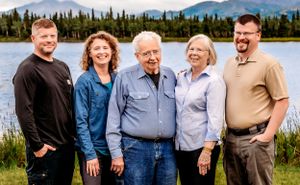 Five people stand together by a lake with mountains and evergreen forest in the background.