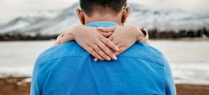 Close-up of hands embracing from behind with snowy mountains in the background.