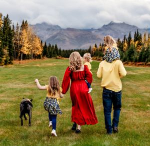 Family walks through mountain meadow with dog against backdrop of autumn colored trees.