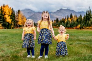 Three children in matching floral outfits laugh together against mountain backdrop with fall colors.