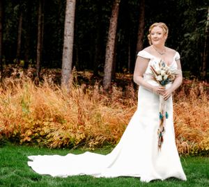 A bride in a flowing white dress holds a bouquet while standing in front of an autumn forest.