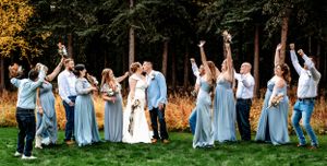 Wedding party celebrates with raised arms on grass with autumn colors in the background.