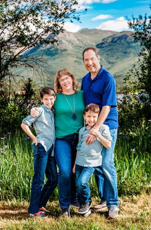 A family poses together outdoors against a scenic mountain backdrop with tall grass and blue sky.