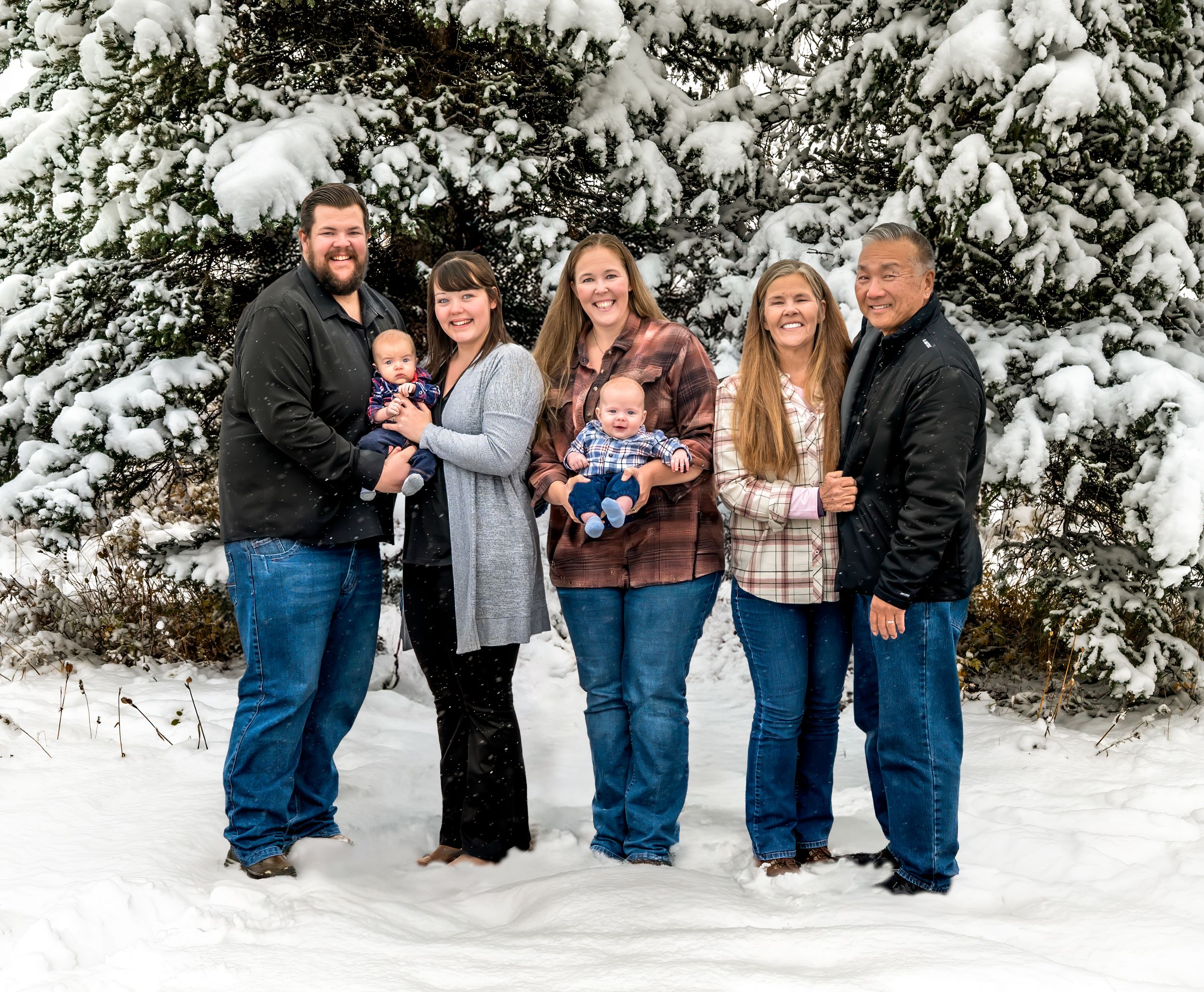 A group of people stand together in a snowy winter setting surrounded by snow-covered evergreen trees.