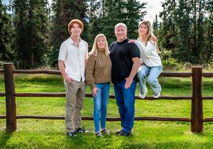 Family members lean against wooden fence posts in a green field surrounded by evergreen trees on a sunny day.