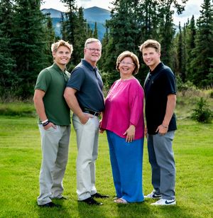 A group of people in casual attire standing together outdoors against a backdrop of evergreen trees and mountains.