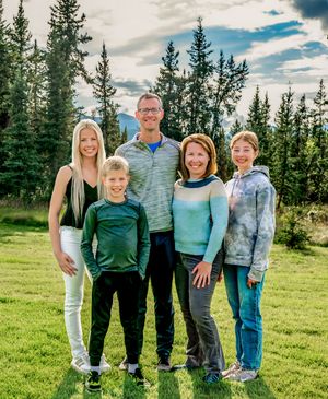 A group in coordinated blue and green outfits poses together on a grassy hill with evergreen trees in the background.