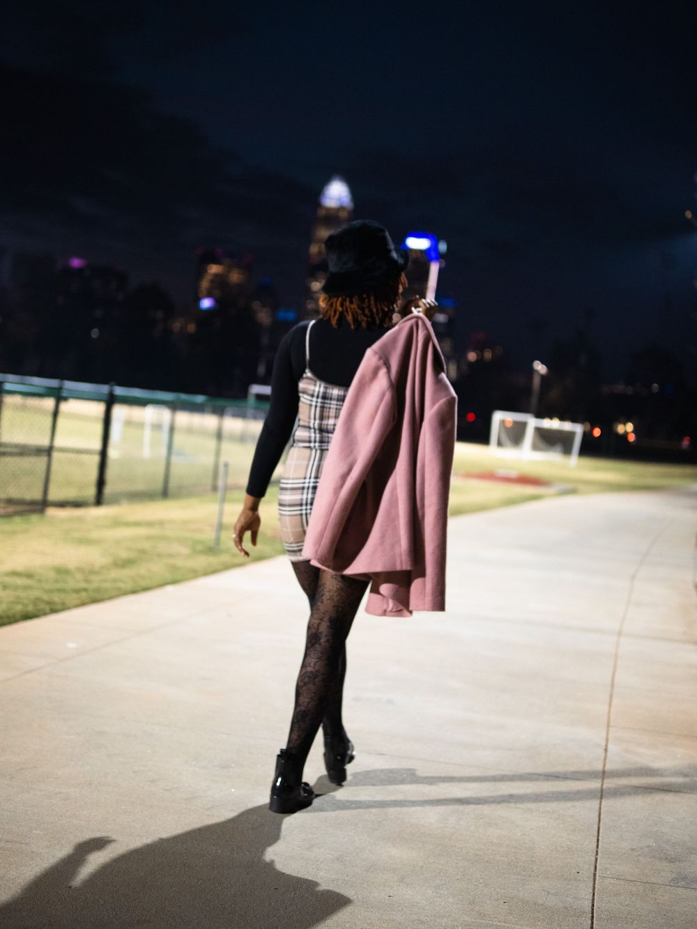 A figure walks on a concrete path at night carrying a pink jacket, wearing a black top and dress in dim lighting.