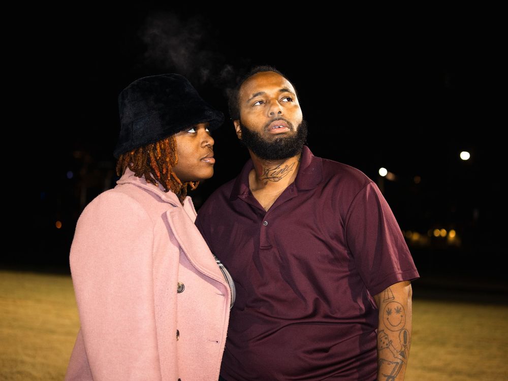 A couple wearing pink coat and burgundy shirt stand together in a dark outdoor setting at night.