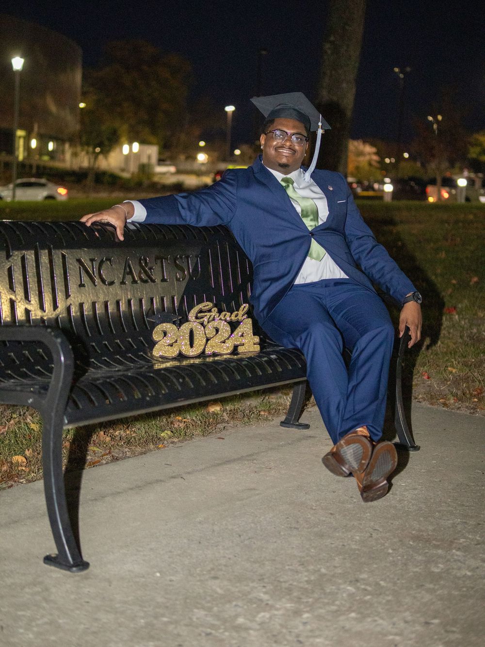 A graduate in a navy blue suit and graduation cap reclines on a decorative bench with '2024' lettering at night.