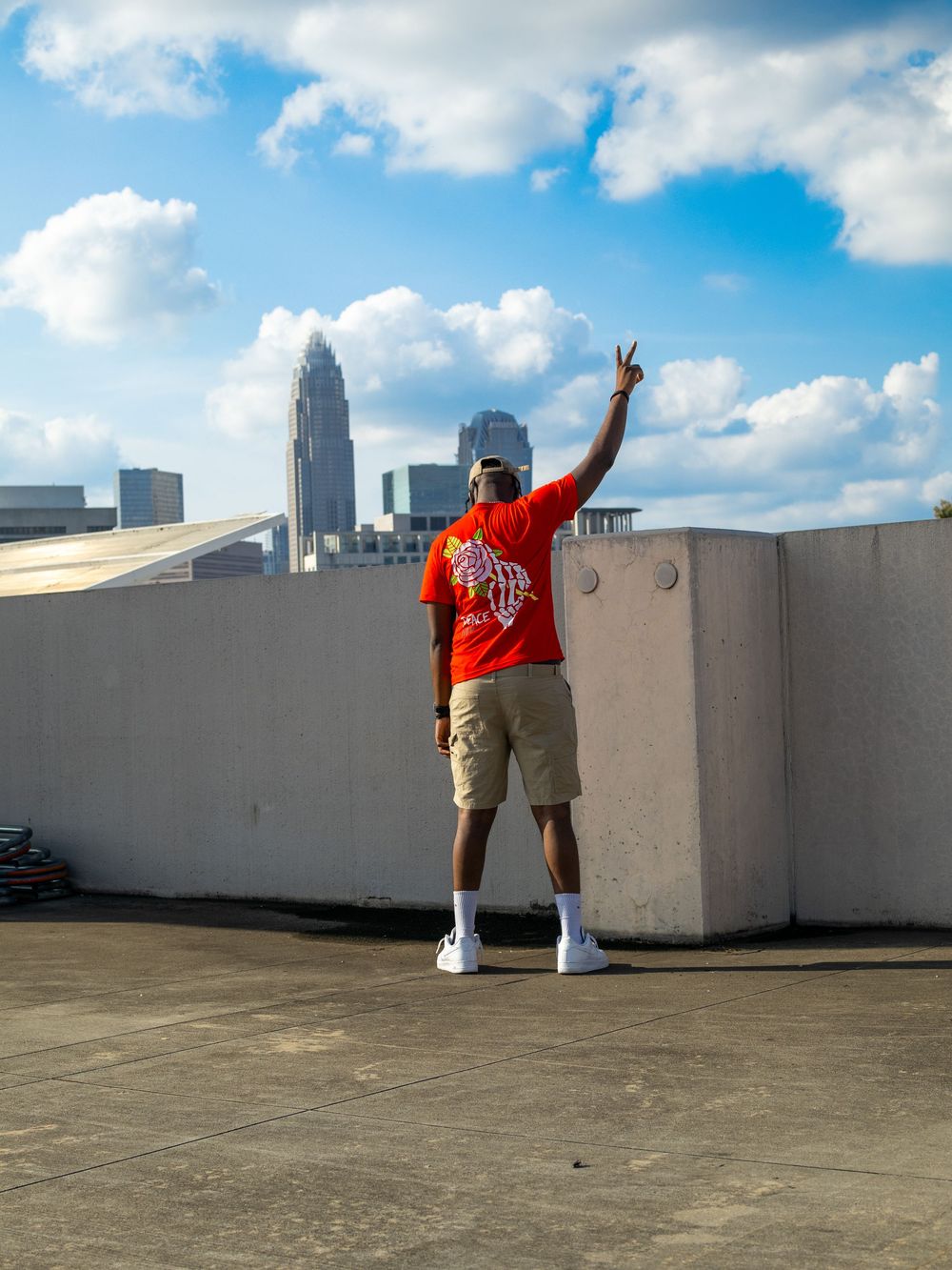 Person in orange t-shirt stands on rooftop making peace sign against Charlotte skyline on sunny day with clouds.