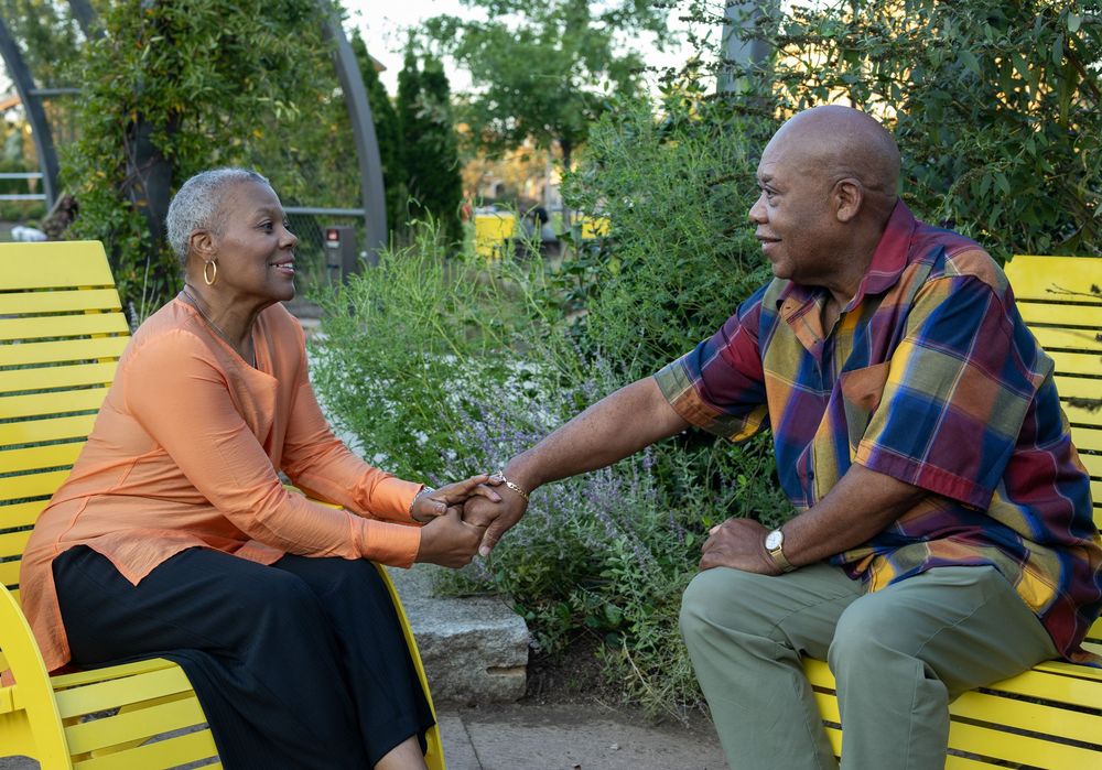 Two seniors sit on yellow park benches having an animated conversation in a garden setting.