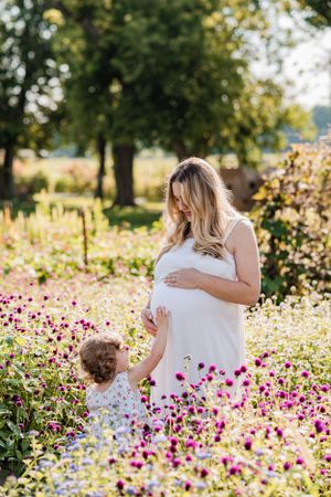 Expecting mother and child standing in field of pink wildflowers on sunny day.