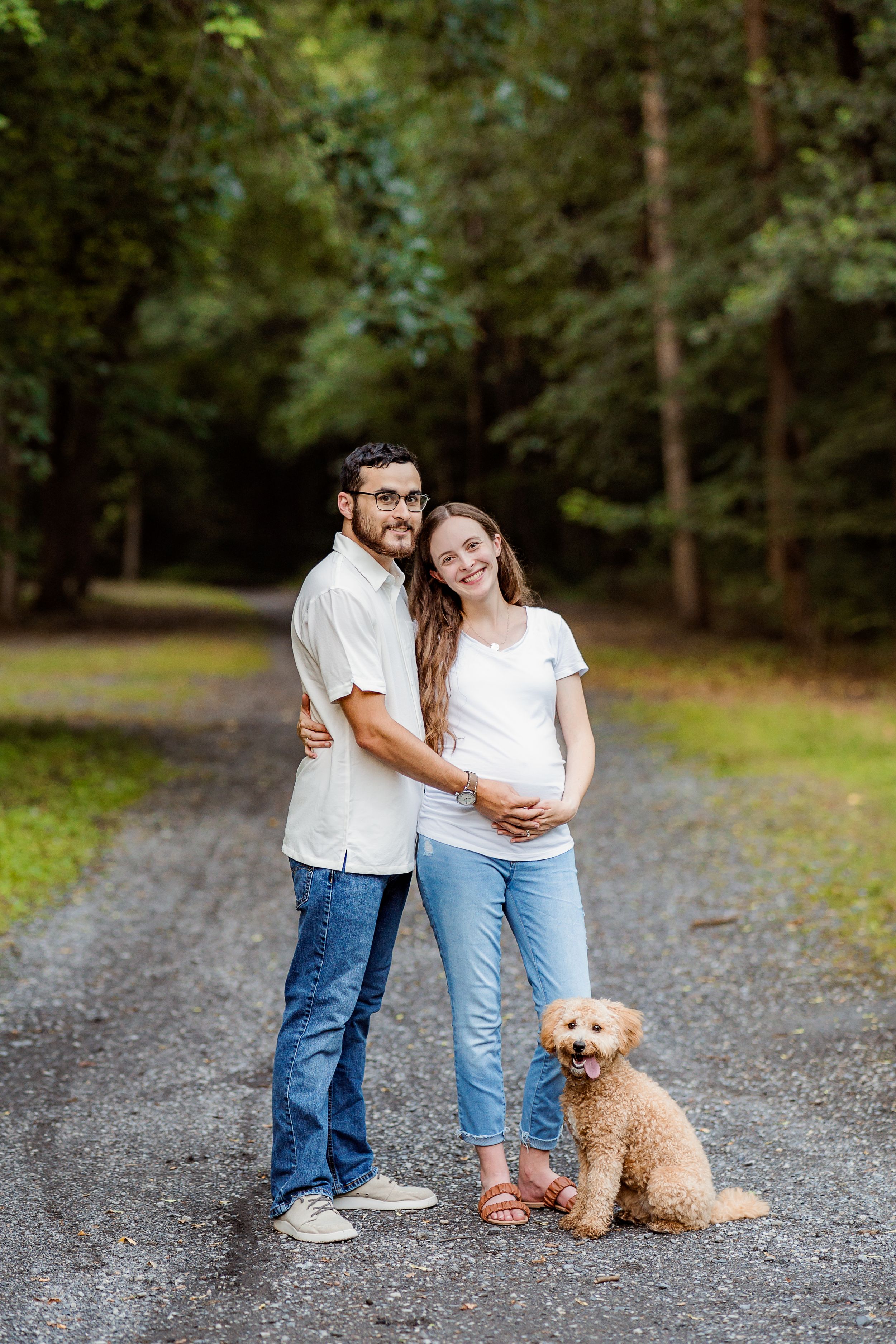 A couple poses with their goldendoodle puppy on a gravel path surrounded by lush green forest.