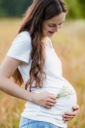 A person in a white t-shirt and jeans holds their pregnant belly while standing in a golden field during sunset.