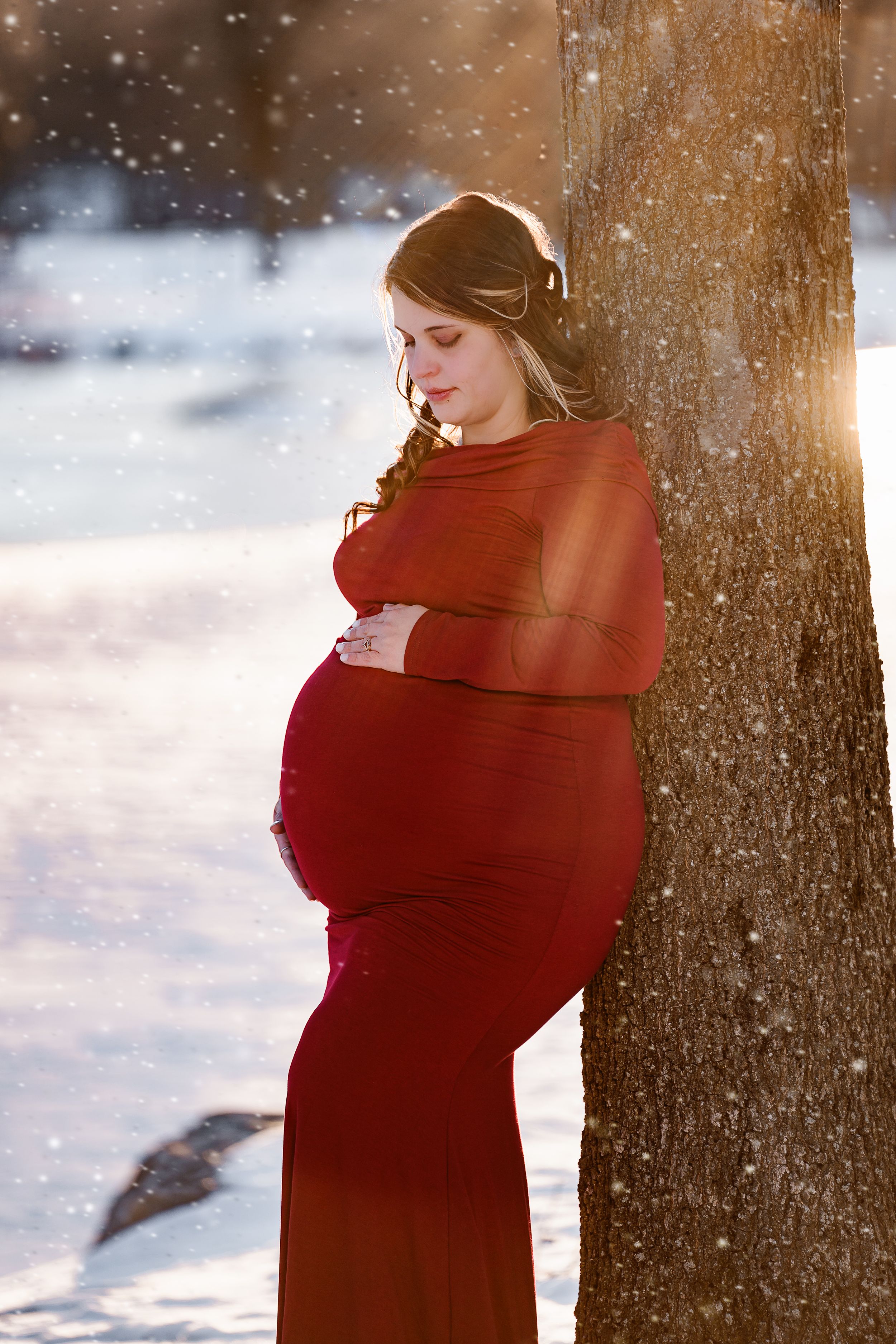 Winter maternity portrait with snow falling and sun flare through trees.
