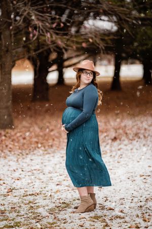 Woman in teal dress poses in snowy winter setting among bare trees wearing a brown hat.