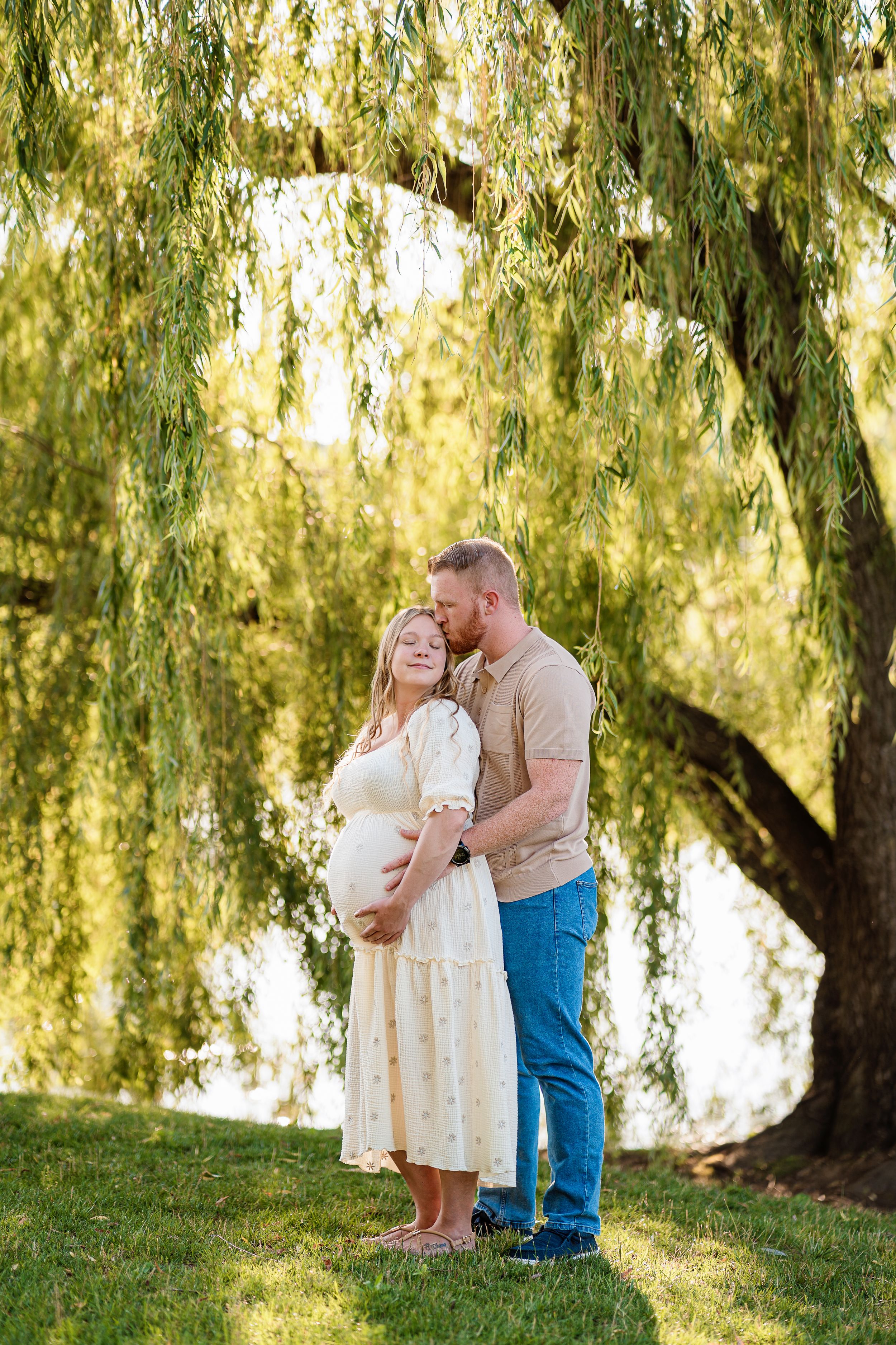 A couple embraces beneath a weeping willow tree during a romantic maternity photography session on a sunny day.