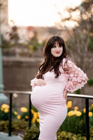 Expectant mother in a pink form-fitting dress with floral sleeves poses outdoors at sunset.