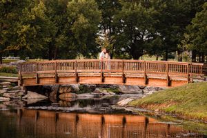 A rustic wooden footbridge spans across a peaceful pond with rocks along the shoreline in an autumn park setting.