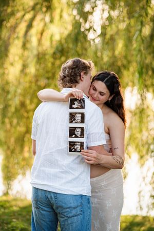 Couple sharing intimate moment outdoors while displaying ultrasound photos on white shirt against willow tree background.