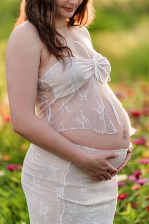 A pregnant woman in a white lace dress poses in a garden with pink flowers at golden hour.