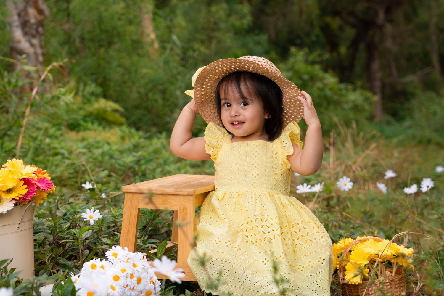Adorable toddler girl in yellow dress and straw hat posing among colorful flowers outdoors.