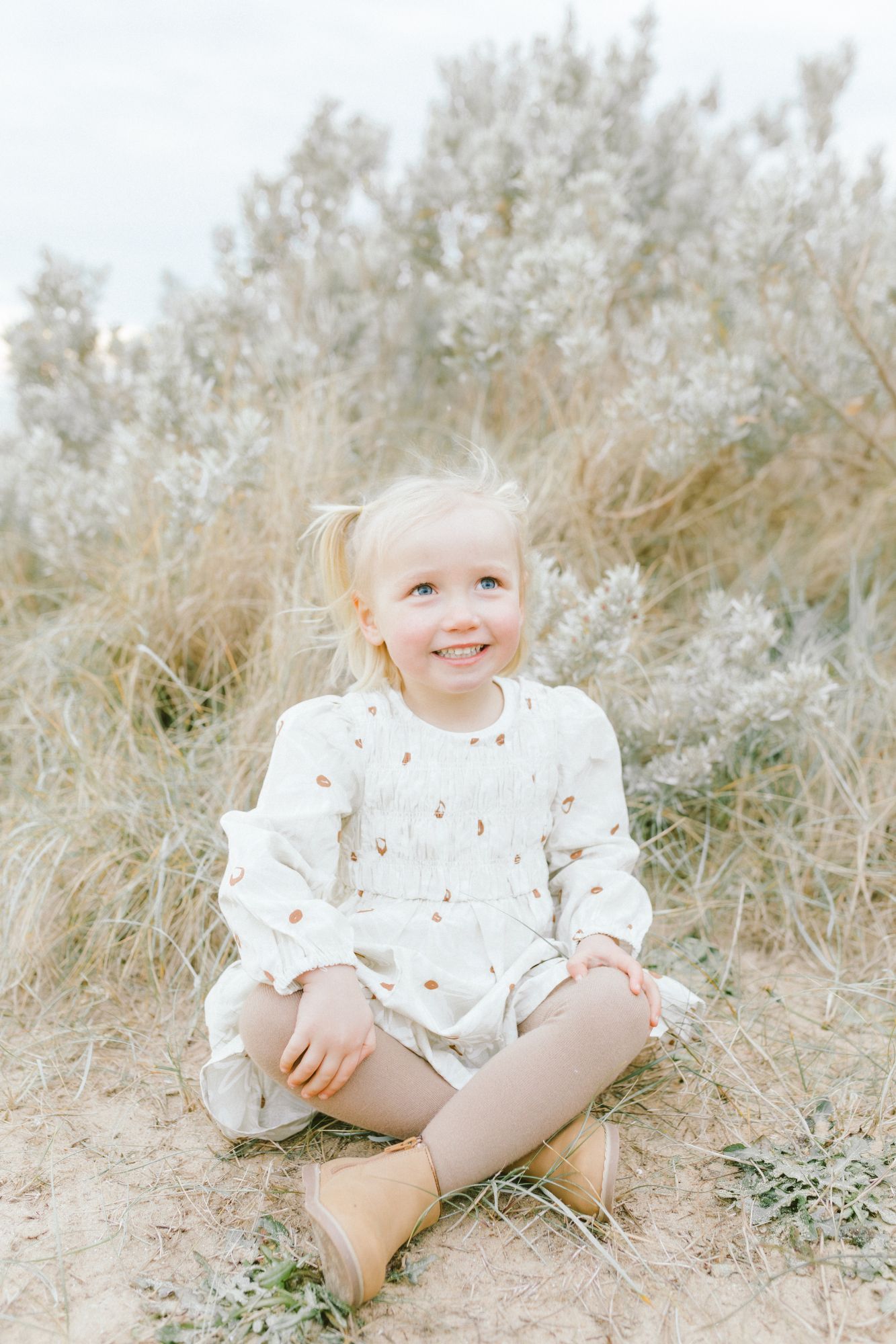 A small child in a white floral dress sits among tall dried grasses in a dreamy outdoor setting.