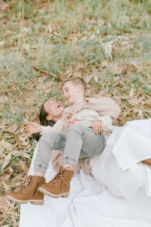 Two people laying on a white blanket in autumn leaves wearing casual clothing and brown boots.