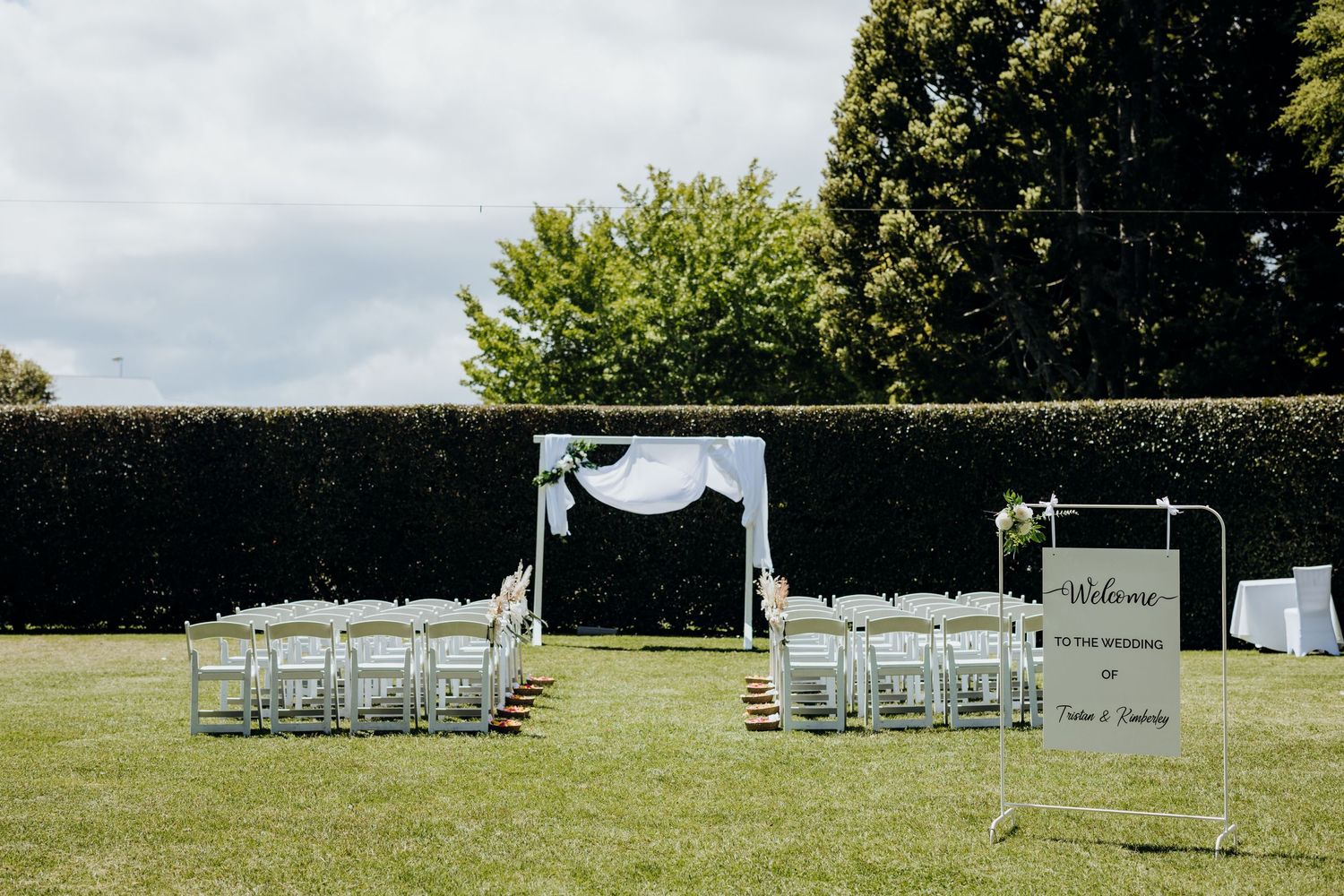 Outdoor wedding ceremony setup with white chairs and an arch draped in fabric on a manicured lawn with hedges.