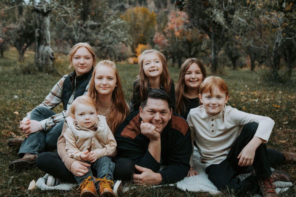 A father lies on a blanket surrounded by his smiling children and a baby during an autumn family photo session in an orchard.