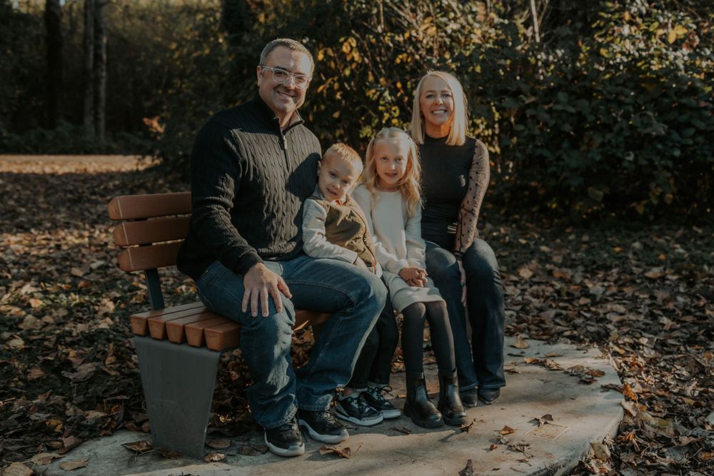 A smiling family of four sits together on a wooden park bench surrounded by fallen autumn leaves in a cozy outdoor portrait.