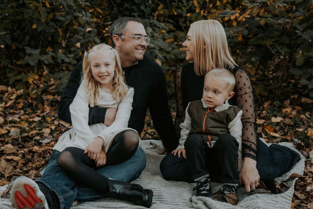 Smiling family of four sits together on a blanket covered in autumn leaves during an outdoor fall family portrait session.