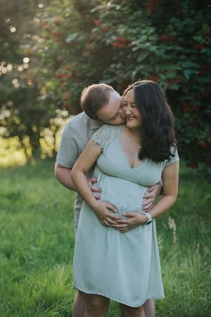 Familienfotografie: Schwangeres Paar steht im Grünen, der Partner hält die Hände schützend um den Babybauch.