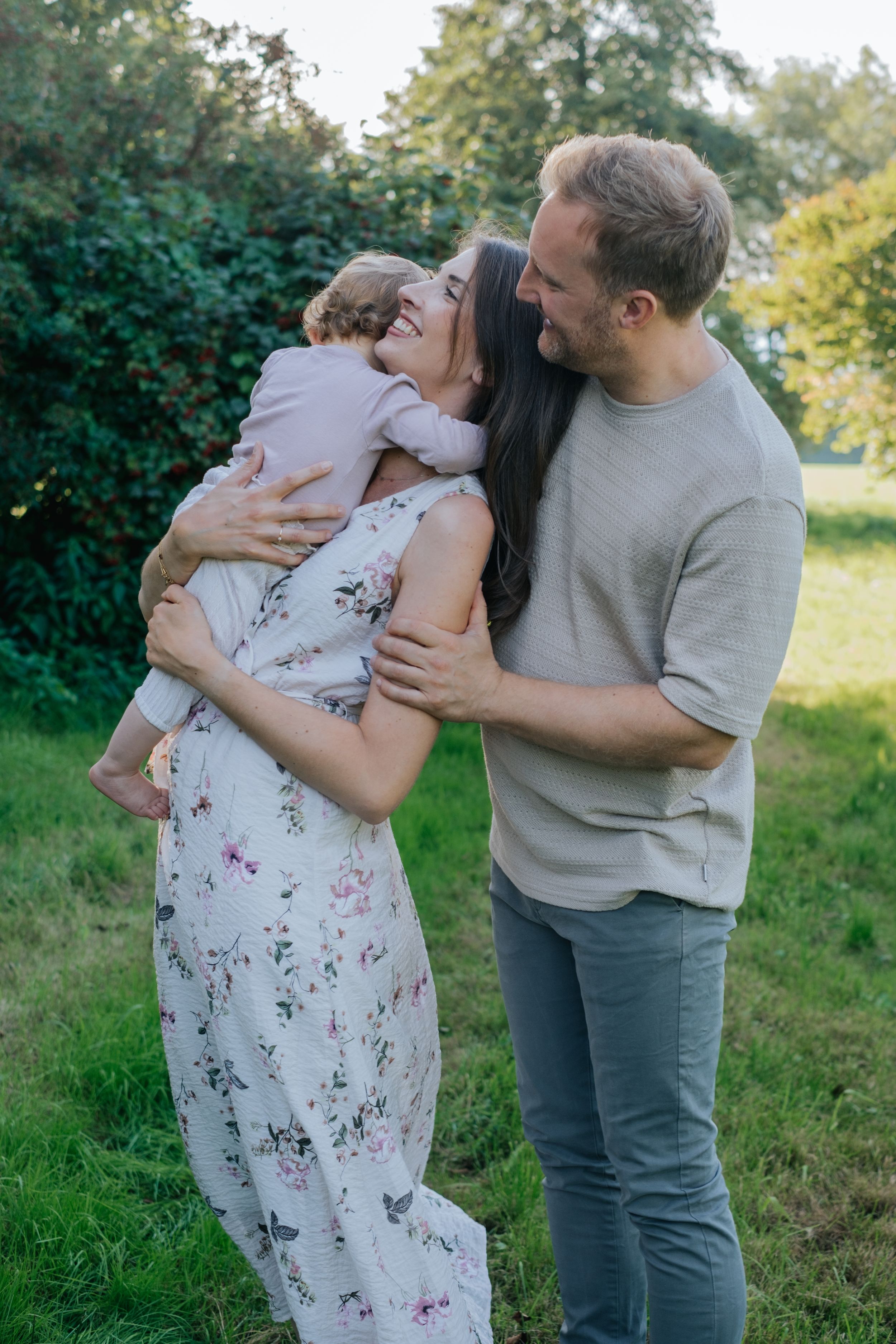 Familienfotografie: Eltern stehen im Grünen und halten ihr Baby im Arm, natürliche Aufnahme im Alltag.