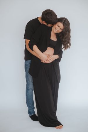 Familienfotografie: Schwangeres Paar steht eng umarmt vor hellem Hintergrund, ruhige Studioaufnahme.