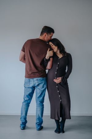 Familienfotografie: Paar steht eng beieinander vor hellem Hintergrund, der Babybauch ist sichtbar.