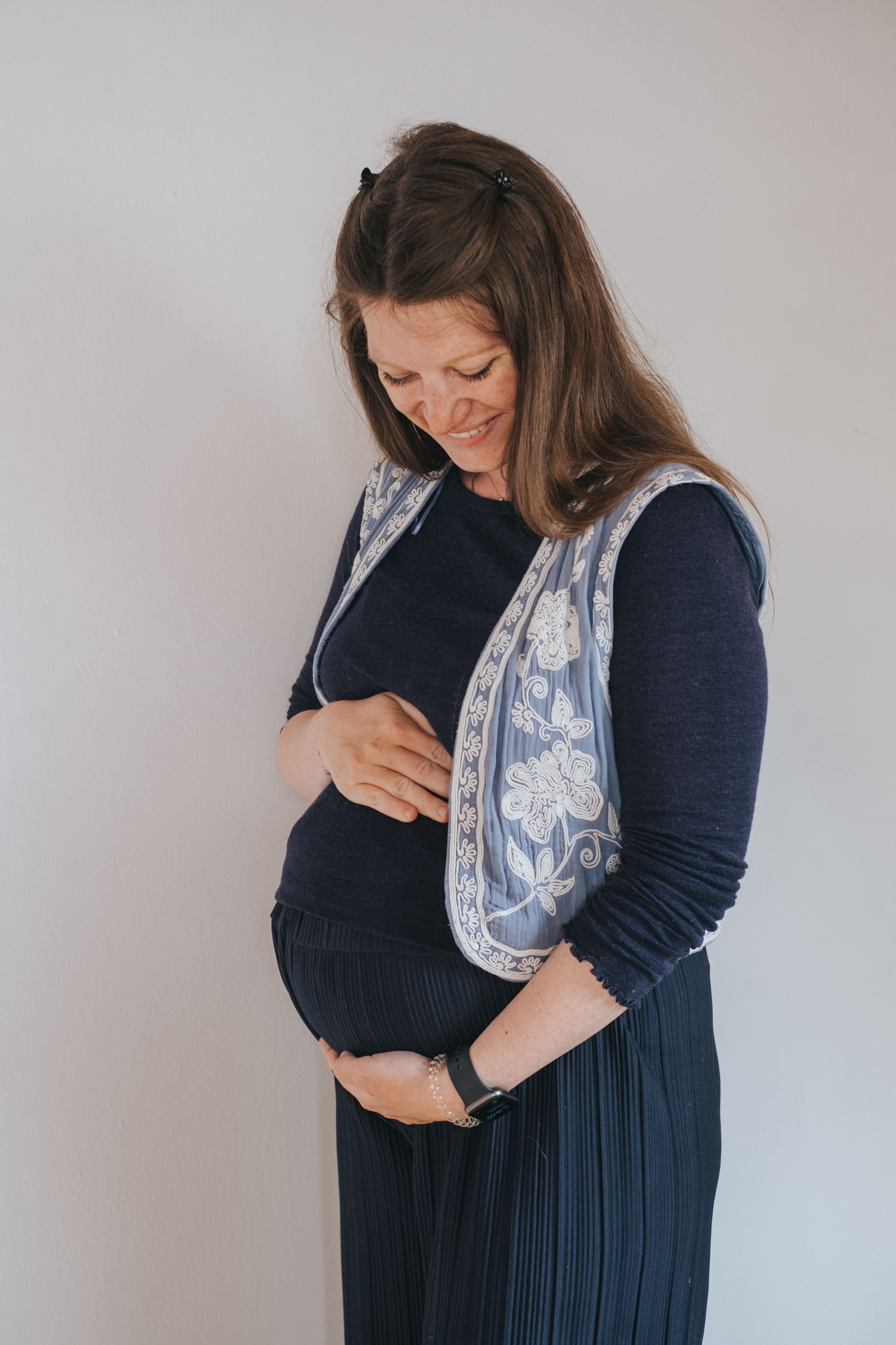 Familienfotografie: Schwangere Frau steht ruhig vor hellem Hintergrund und hält sanft ihren Babybauch.
