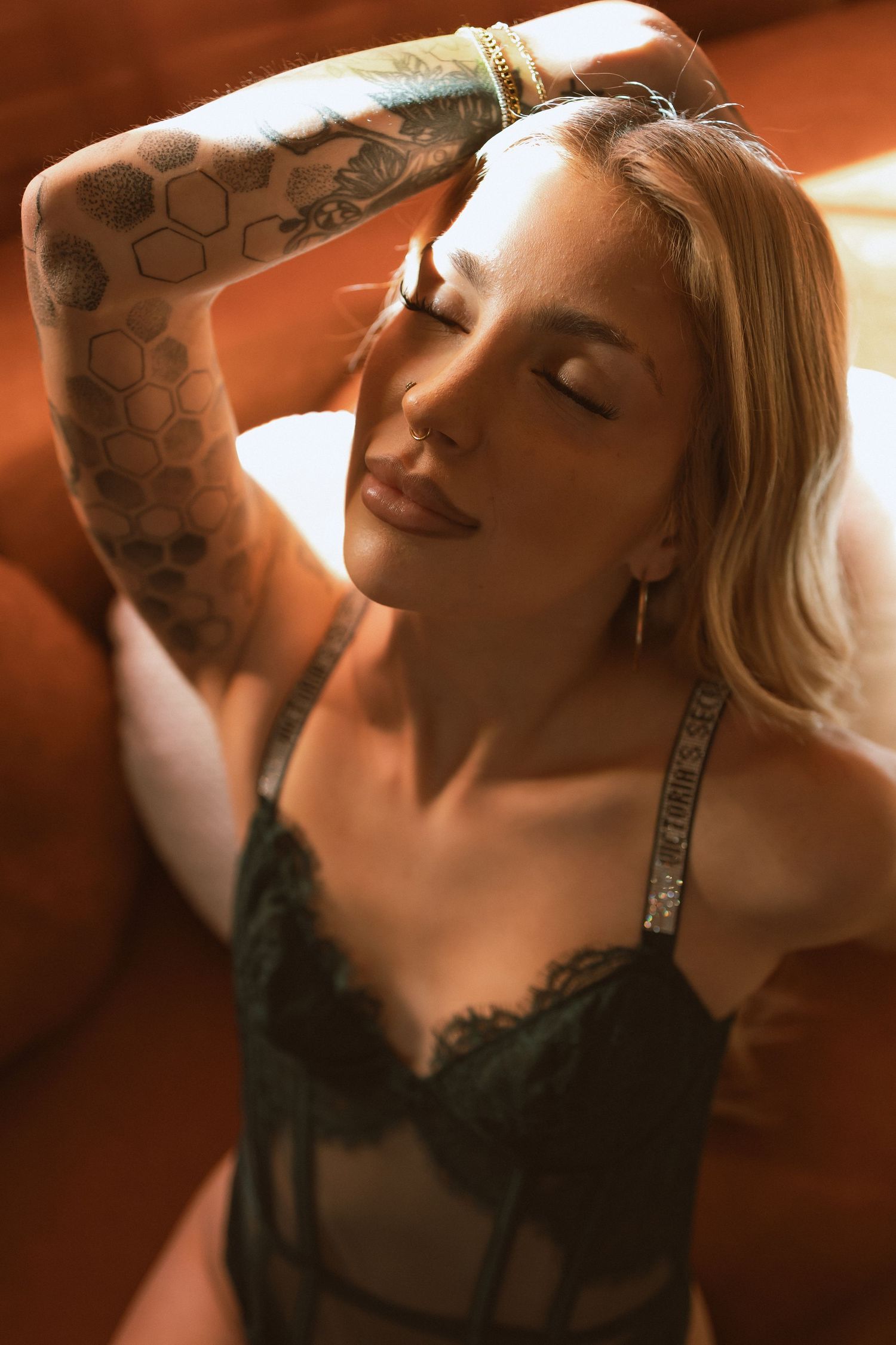 Artistic portrait in warm lighting shows a person with blonde hair and intricate arm tattoos wearing black lace lingerie.