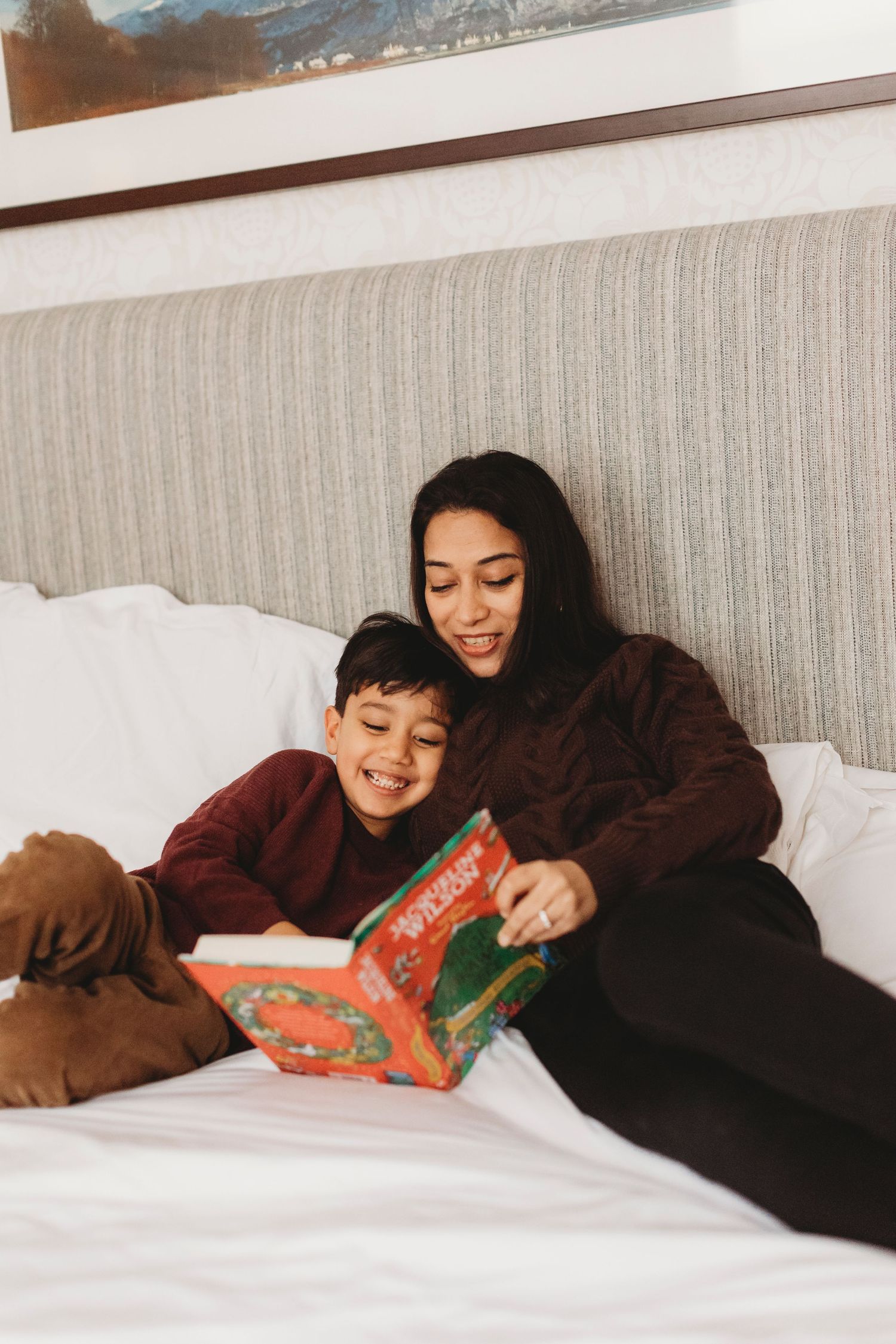 A cozy bedtime reading session shows a parent and child enjoying a storybook together while relaxing on white bedding.