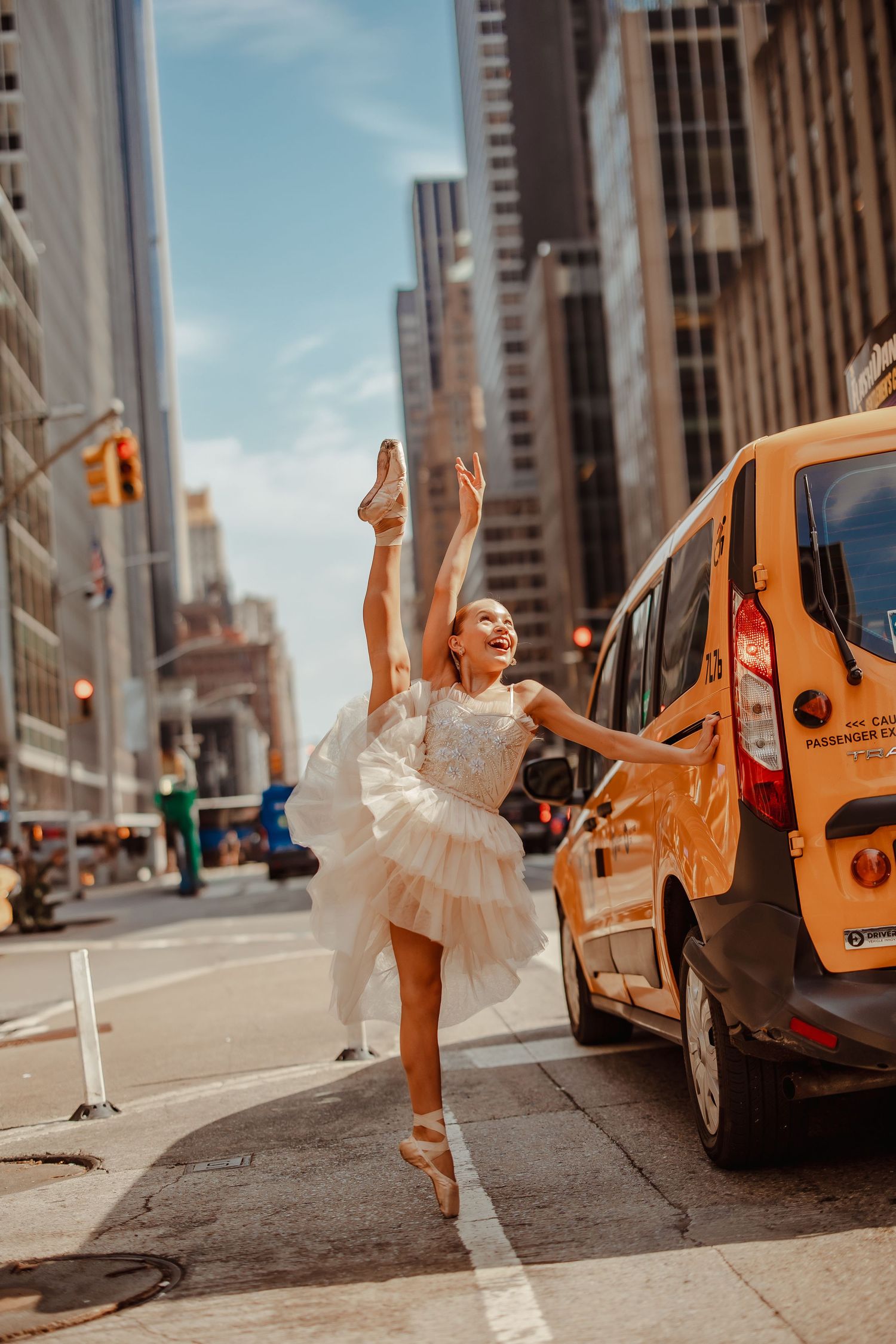 Ballet dancer in white tutu performs elegant pose next to yellow taxi on New York City street.