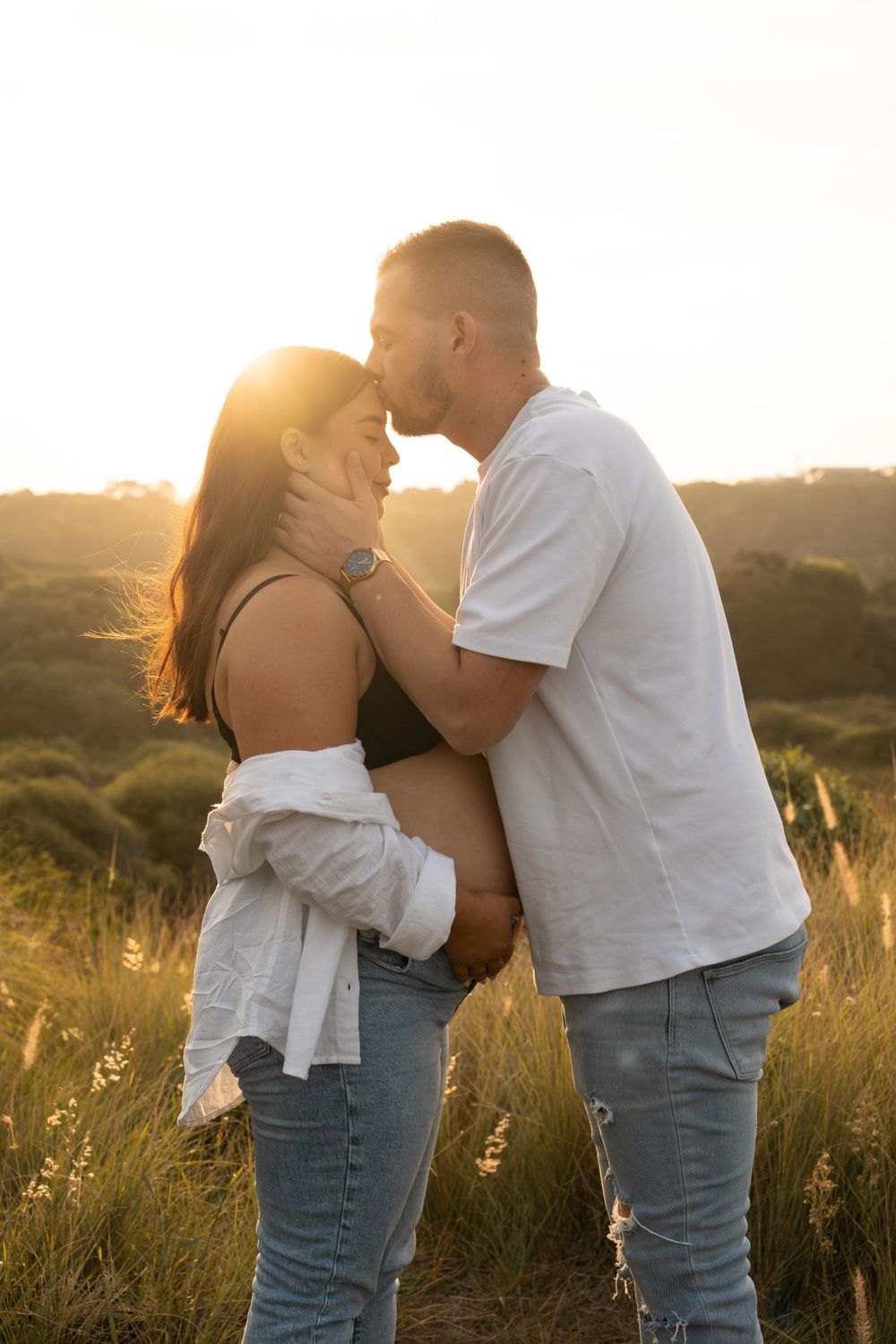 Mother to be holding her pregnant belly while husband kisses her head.