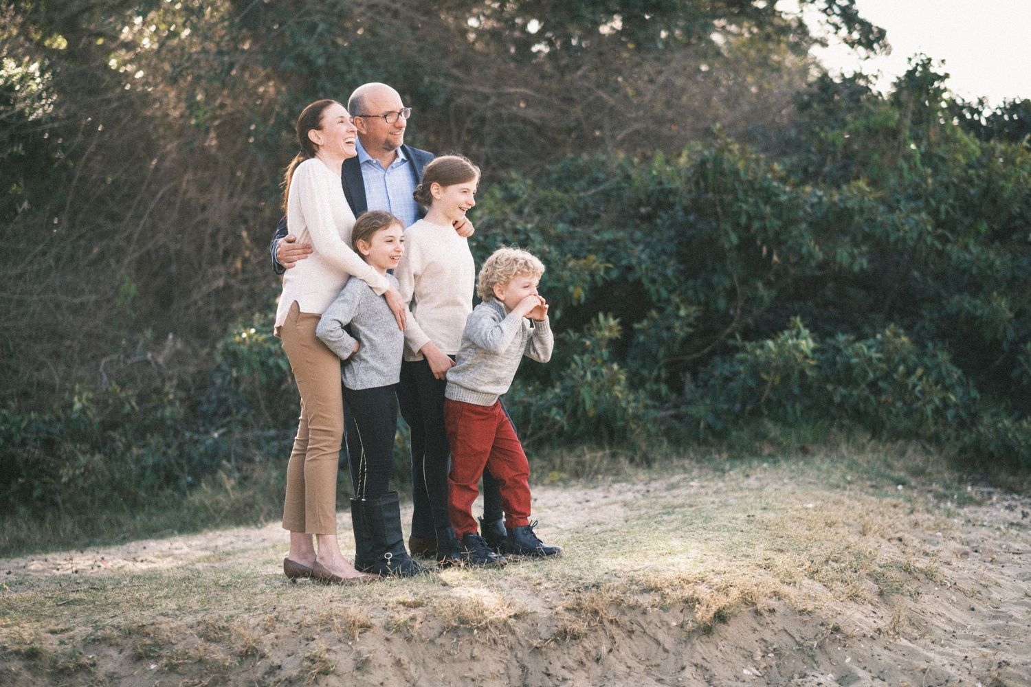Family of five posing outdoors on sandy ground with lush green trees in the background during golden hour.