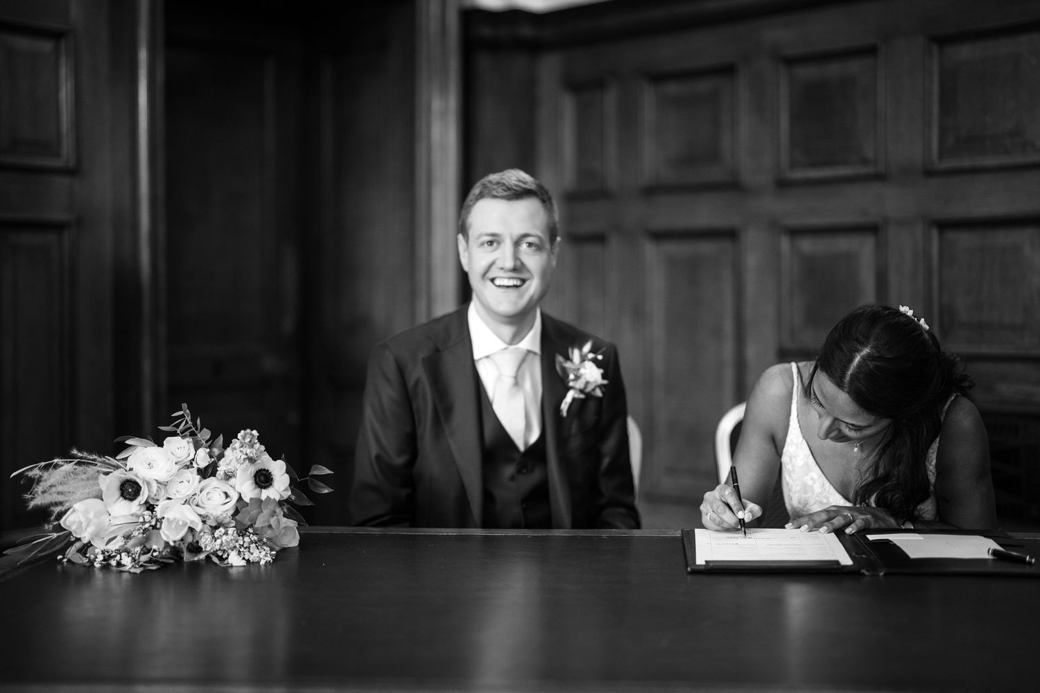 Black and white wedding photo showing a happy moment during marriage registration with flowers on the table.