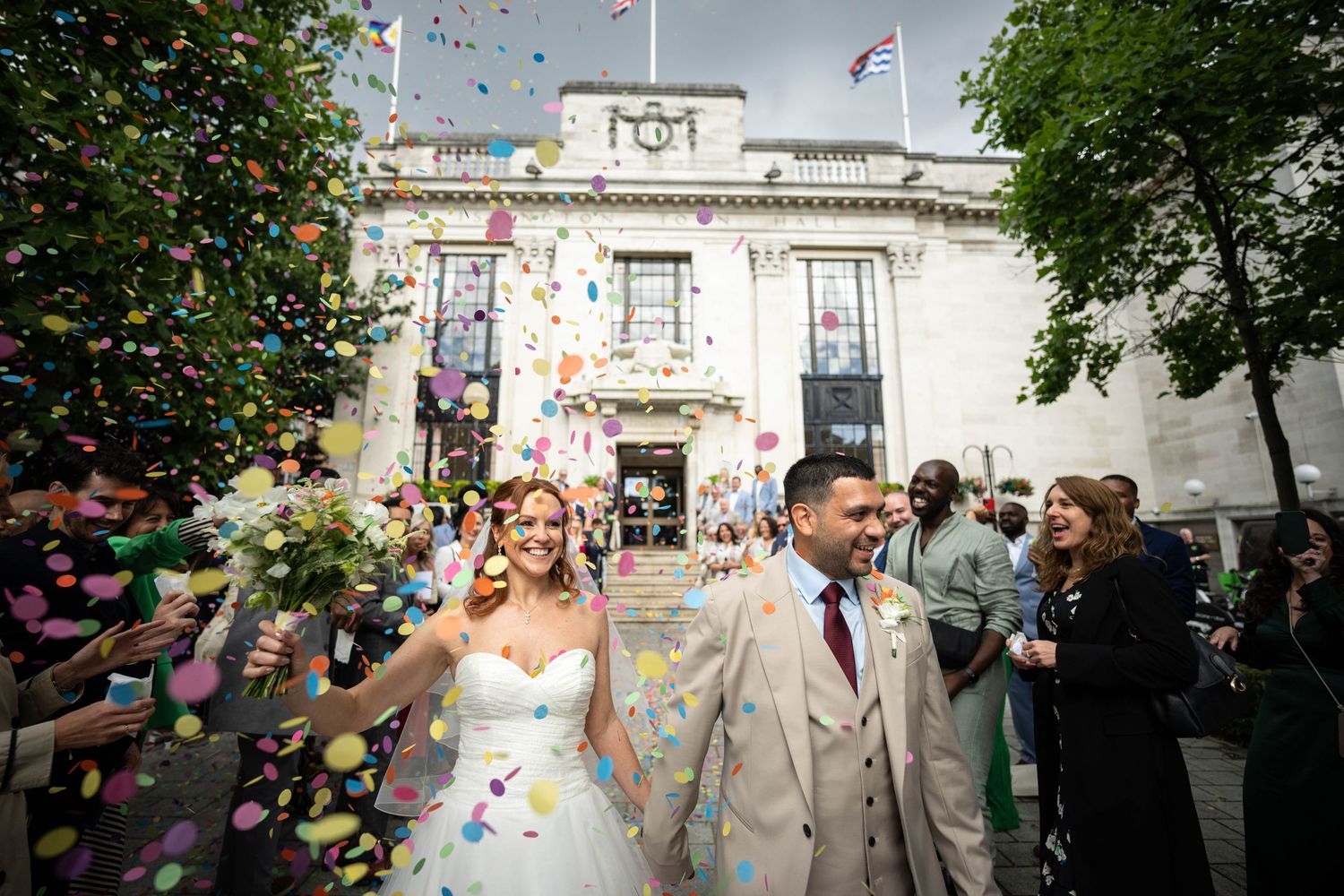 Newlyweds exit a grand building as colorful confetti rains down around them on their wedding day.